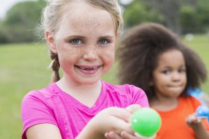 A little girl with freckles, 7 years old, having fun competing in an egg spoon race.  She is smiling at the camera, balancing a green egg on her spoon, friends out of focus in the background.